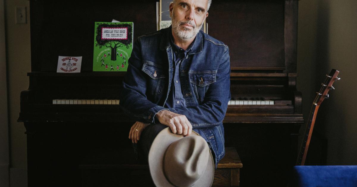 Man with short white beard in jean jacket an holding white cowboy hat leans against an upright piano
